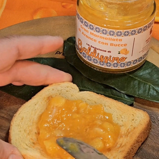 Marmellata di arance con bucce Baldissimo spalmata su una fetta di pane, con vasetto sullo sfondo.